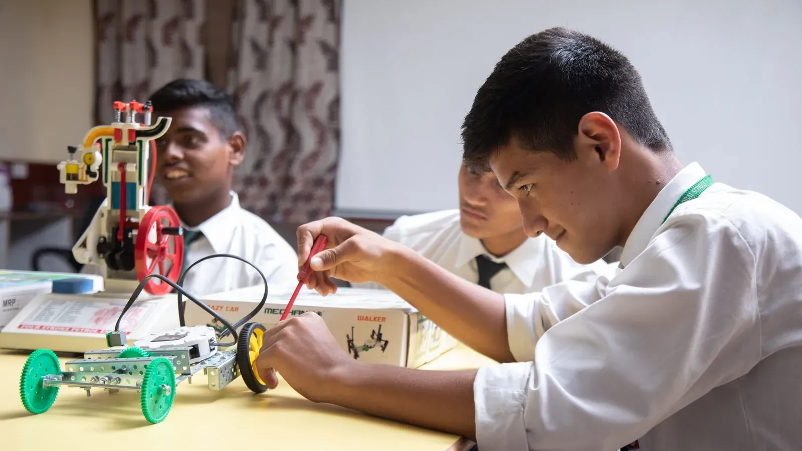 Doon Global School students building a robot car from a construction kit, with a model engine on the desk beside them