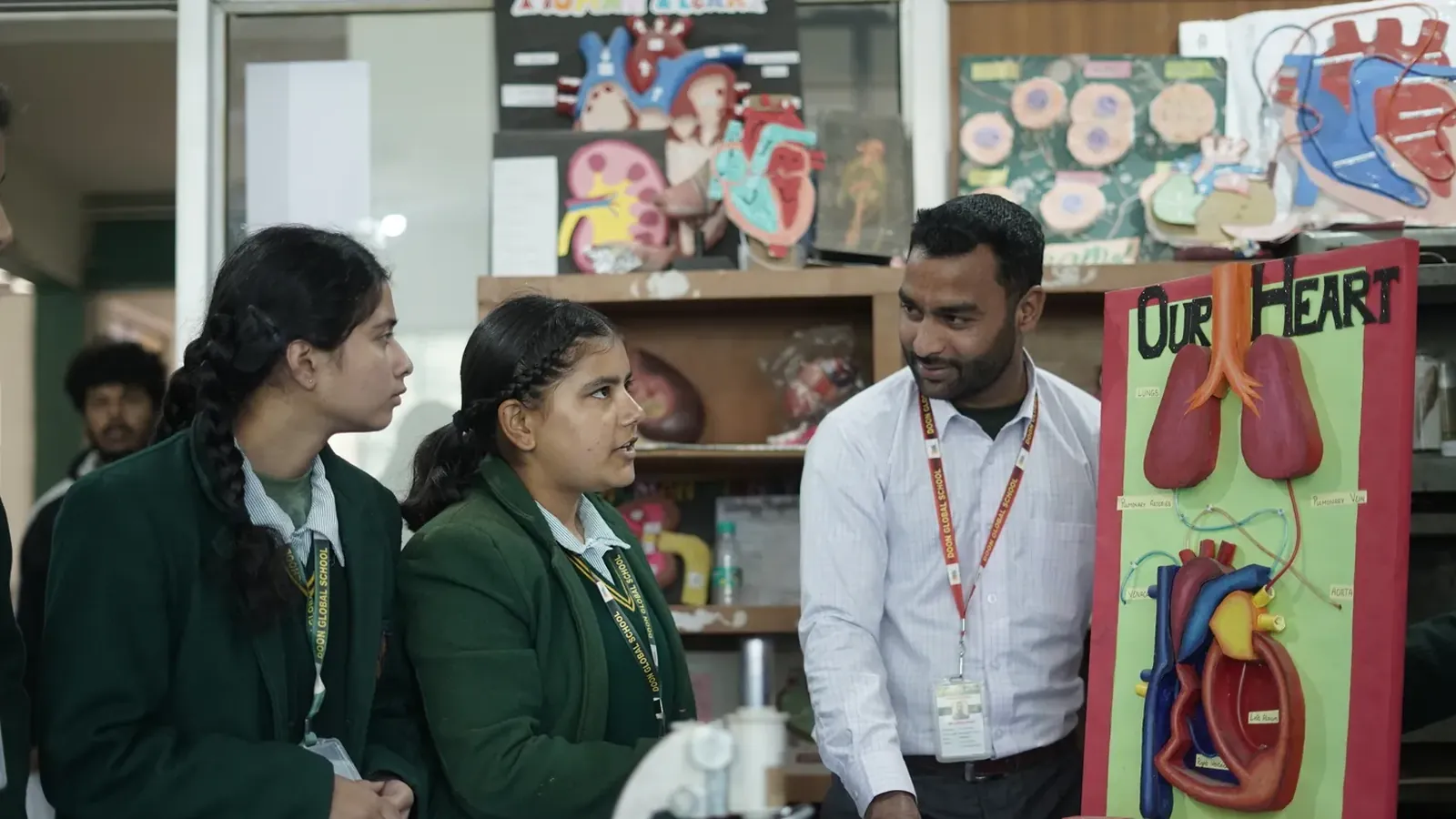 A Doon Global School teacher and two students beside a model of the human heart in the biology laboratory