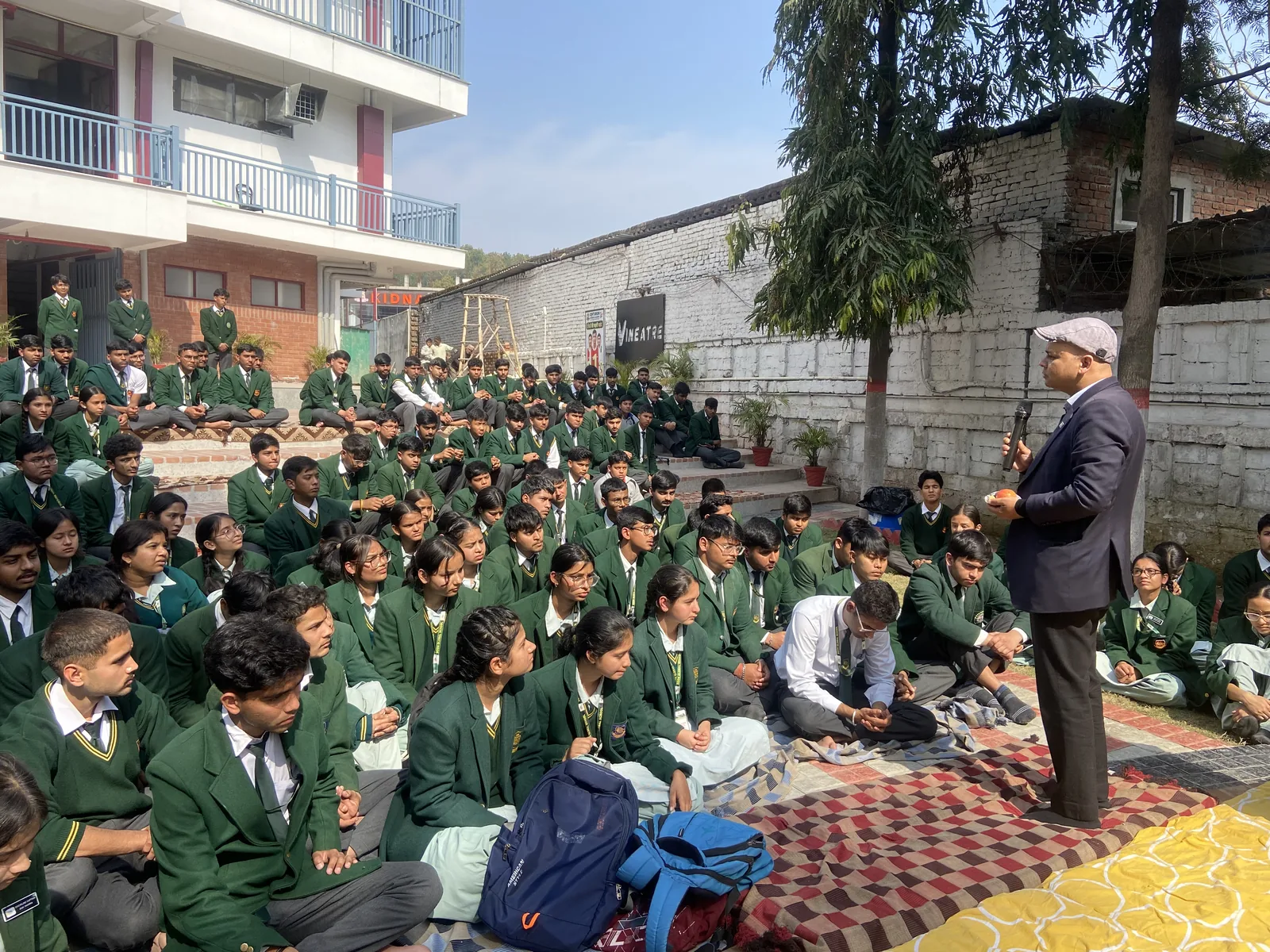 Students seated on the lawn for the Ashirwad ceremony while a teacher addresses them