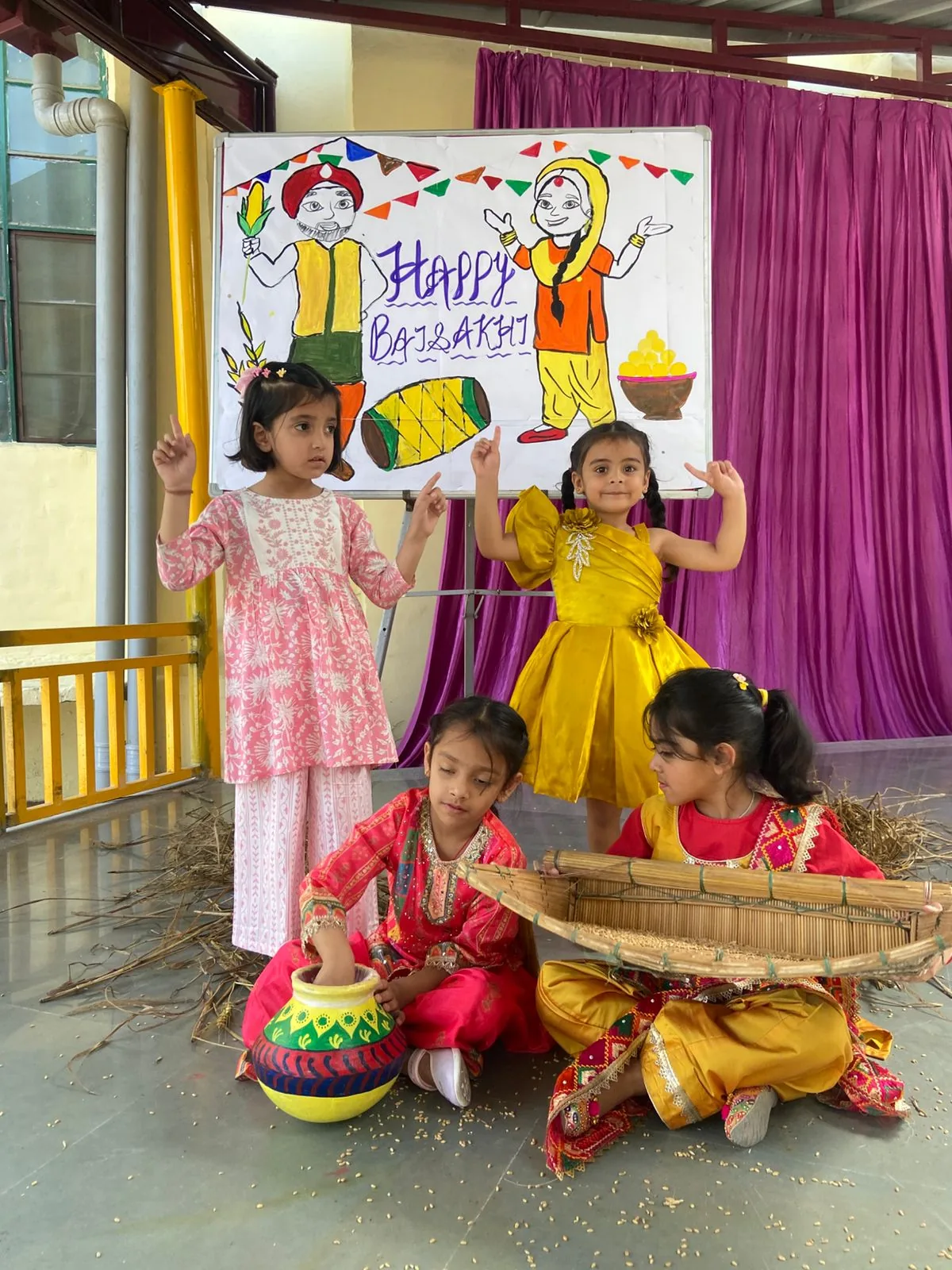 Students in yellow and pink traditional outfits with a Happy Baisakhi poster and a wheat basket